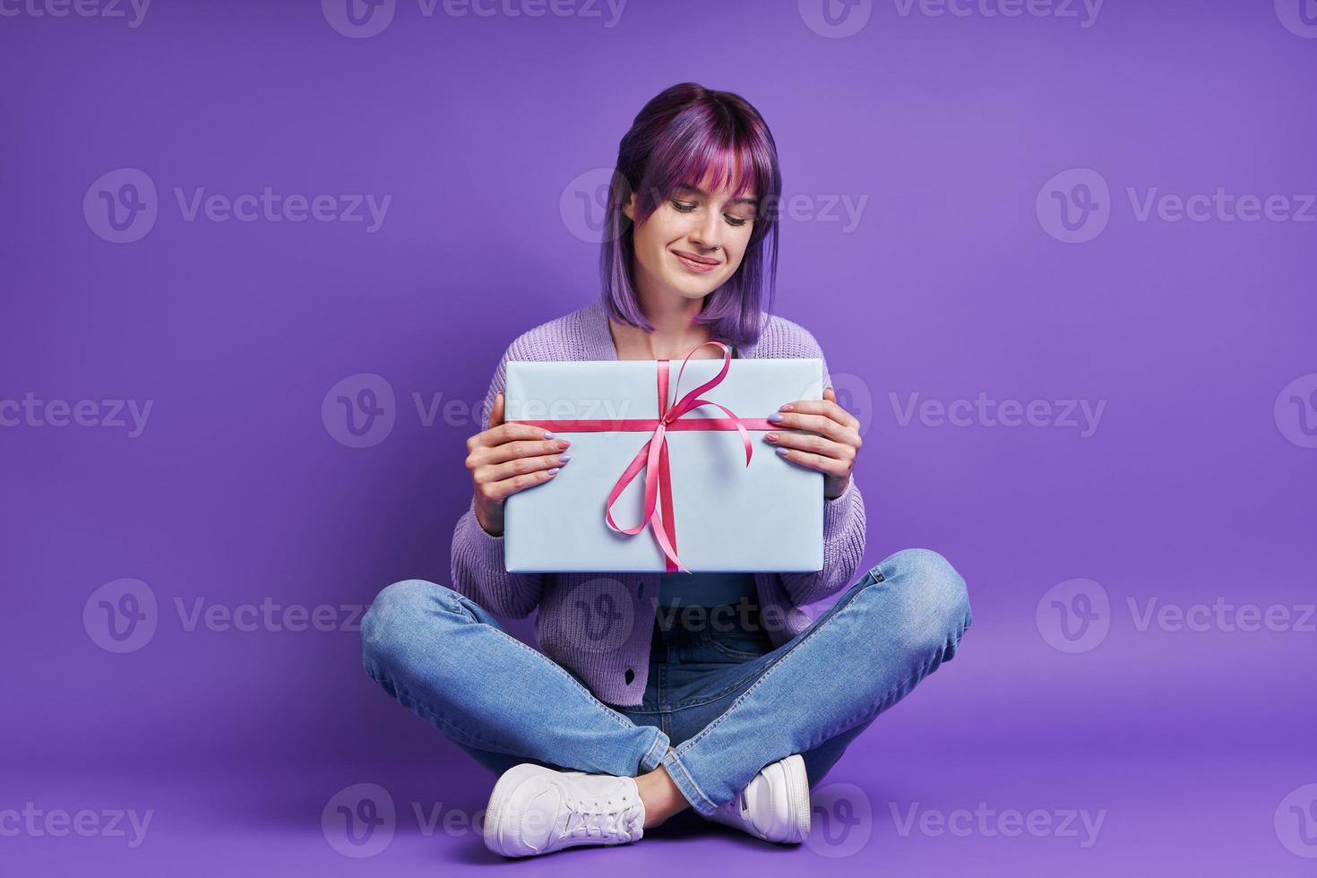 mooi jong vrouw Holding geschenk doos en glimlachen terwijl zittend tegen Purper achtergrond foto