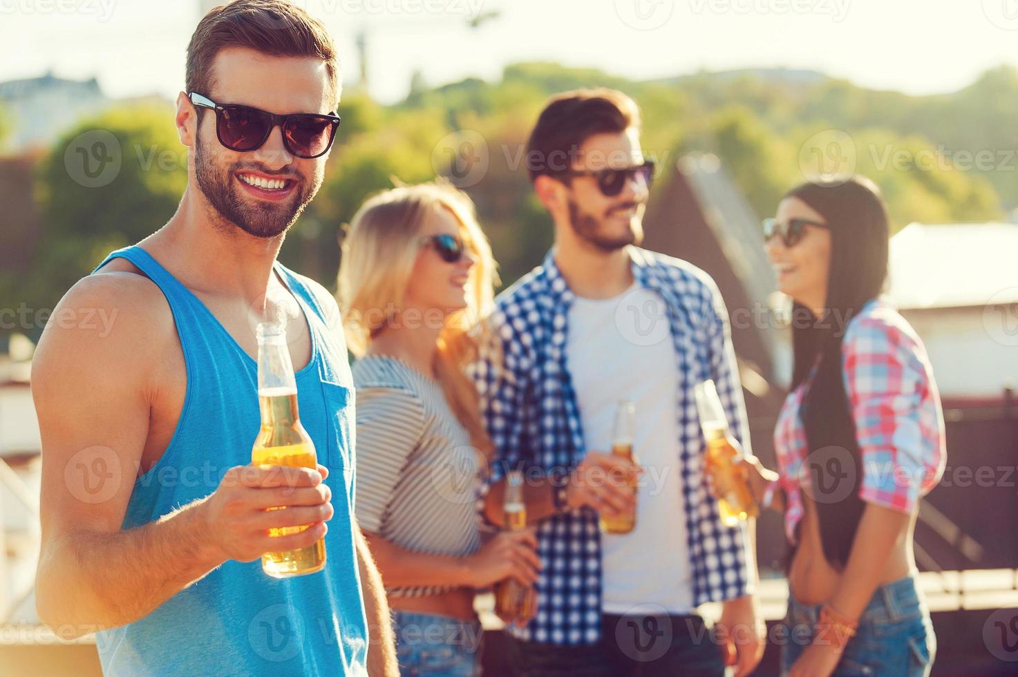 proost gelukkig jong Mens Holding fles van bier en op zoek Bij camera terwijl drie mensen hebben pret in de achtergrond foto