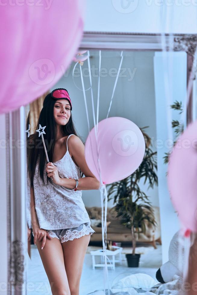 zetten een spellen Aan jij. reflectie van jong aantrekkelijk vrouw in nacht slijtage en slaap masker Holding een magie toverstaf en glimlachen terwijl staand in voorkant van de spiegel foto