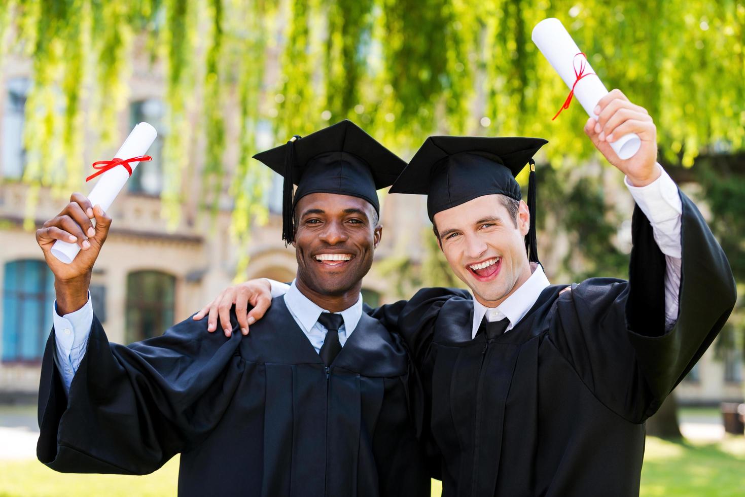 afgestudeerd samen. twee gelukkig jong mannen in diploma uitreiking jurken Holding diploma's en stijgende lijn armen omhoog foto