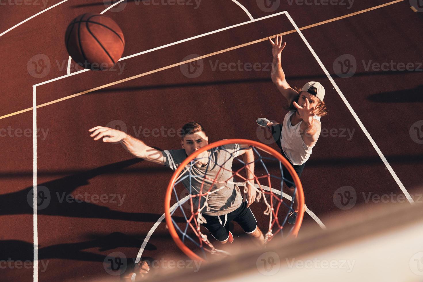 twee jong mannen in sport- kleding spelen basketbal en glimlachen terwijl uitgeven tijd buitenshuis foto