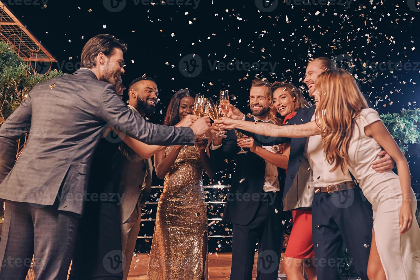 groep van mooi mensen in formele kleding roosteren met Champagne en glimlachen terwijl confetti vliegend in de omgeving van hen foto