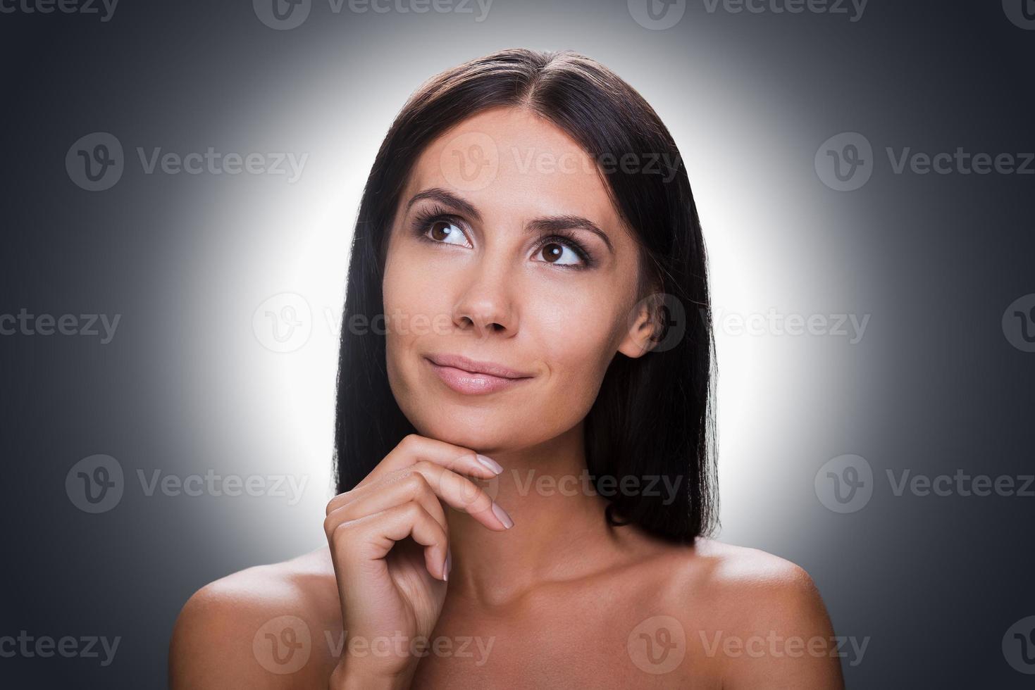 attent schoonheid. portret van attent jong zonder shirt vrouw op zoek weg en Holding hand- Aan kin terwijl staand tegen grijs achtergrond foto