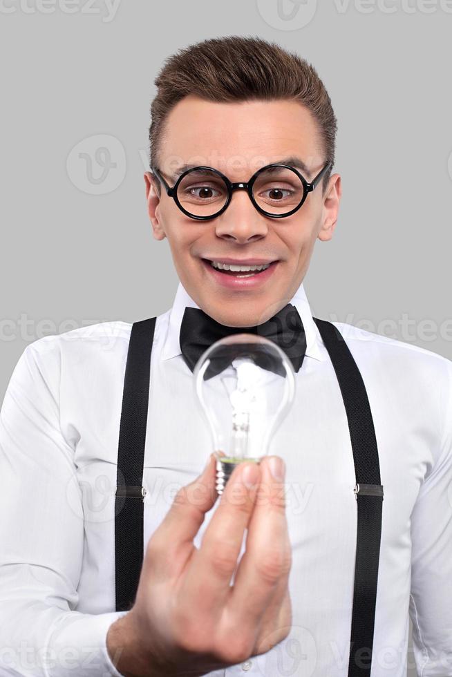 nerd met een licht lamp. verrast jong Mens in boog stropdas en bretels Holding een licht lamp en op zoek Bij het terwijl staand tegen grijs achtergrond foto