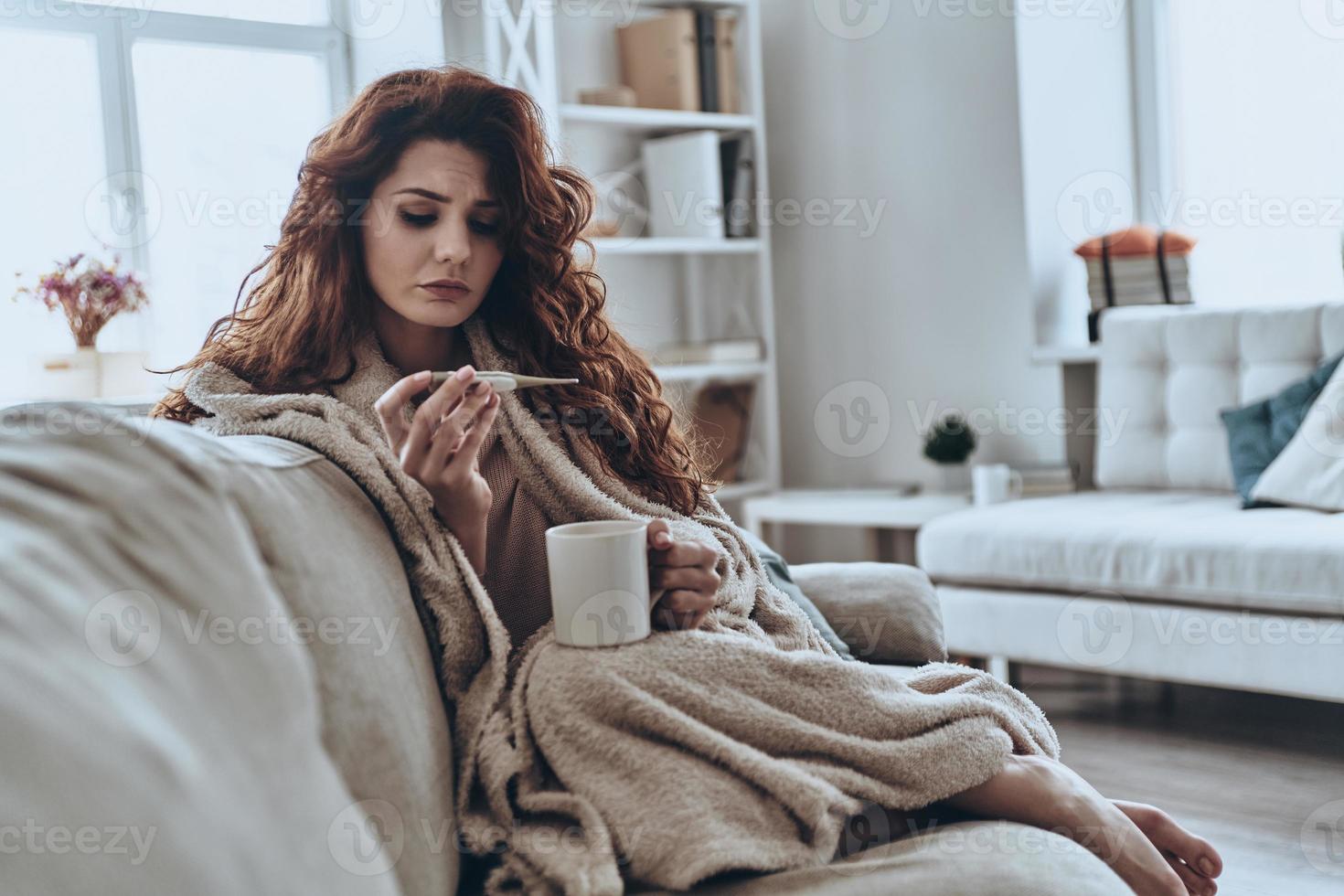 hoog temperatuur. ziek jong Dames op zoek Bij thermometer terwijl zittend Aan de sofa Bij huis foto