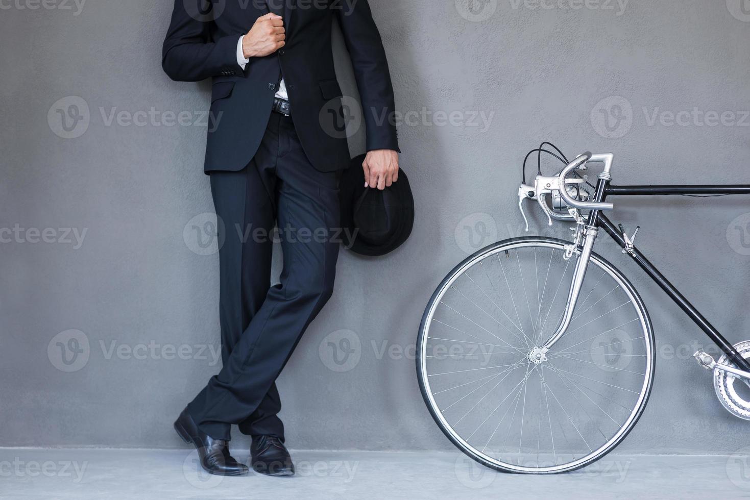 elegant stijl. detailopname van jong zakenmanholding hoed en aanpassen zijn jasje terwijl staand in de buurt zijn fiets tegen grijs achtergrond foto