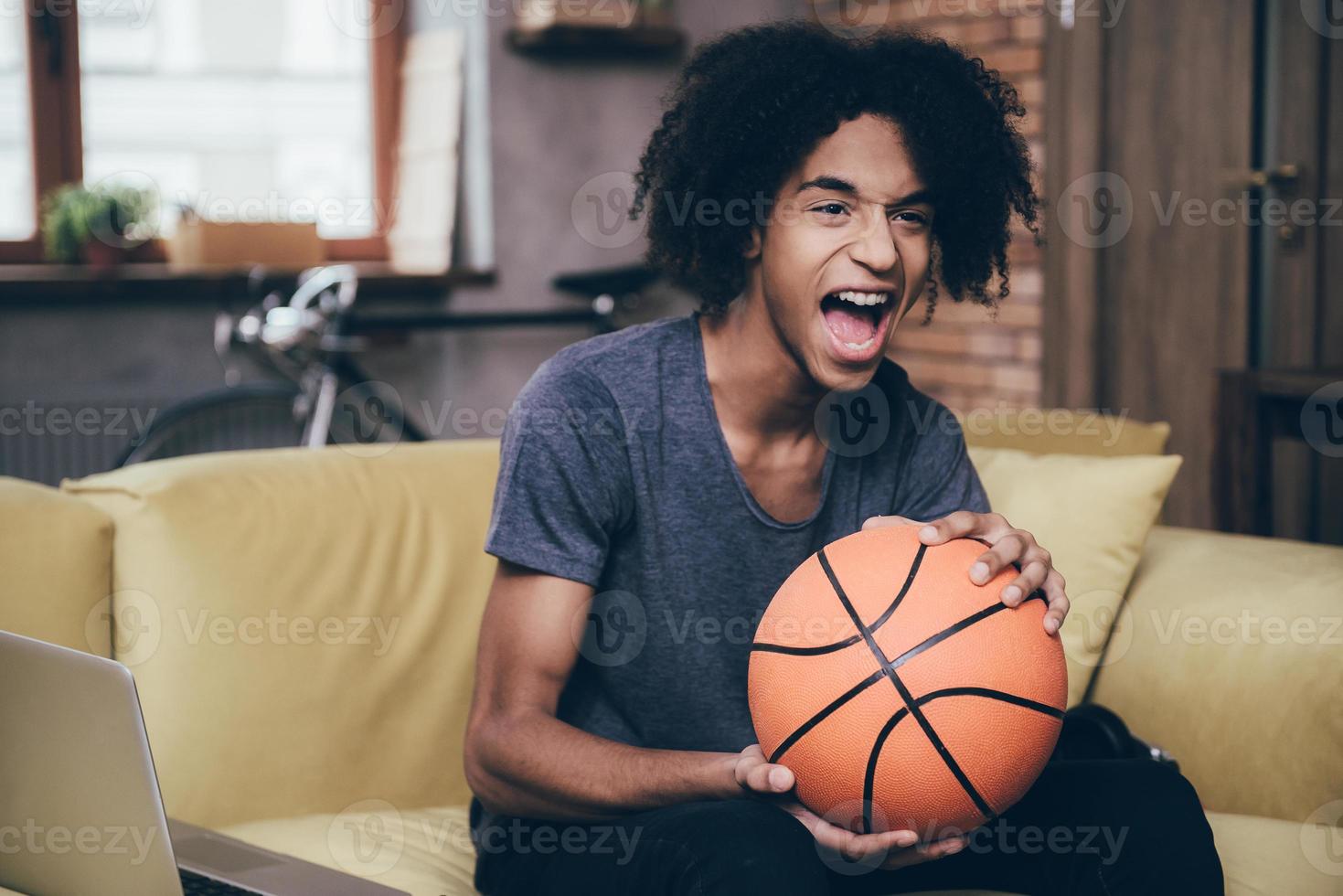juichen voor zijn favoriete basketbal team. vrolijk jong Afrikaanse Mens aan het kijken TV en Holding basketbal bal terwijl zittend Aan de bankstel Bij huis foto