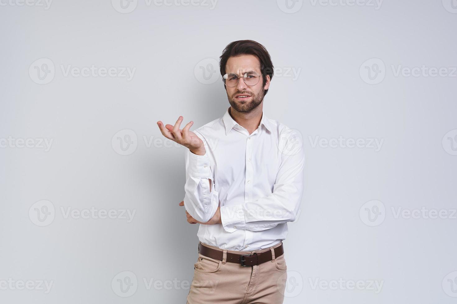 knap jong Mens in wit overhemd op zoek Bij camera en gebaren terwijl staand tegen grijs achtergrond foto