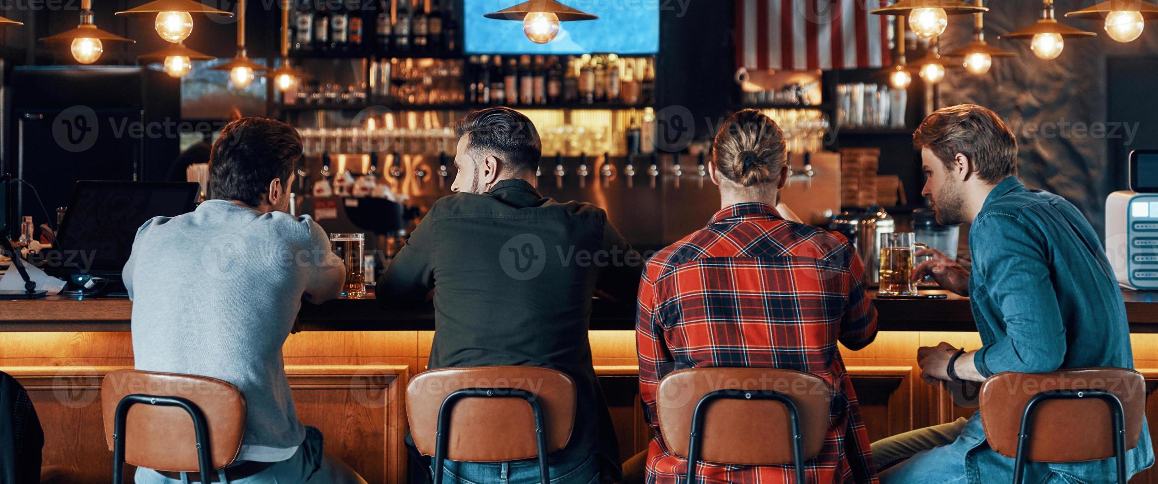 achterzijde visie van jong mannen in gewoontjes kleding drinken bier terwijl zittend in de kroeg foto