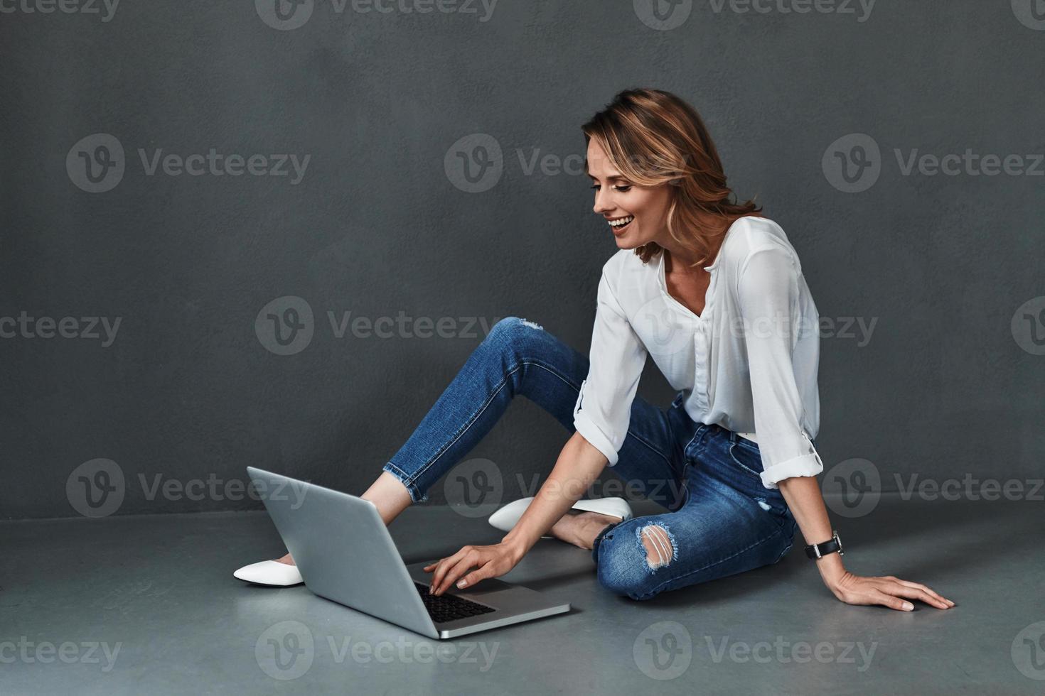 chatten online. aantrekkelijk jong vrouw in gewoontjes slijtage gebruik makend van computer en glimlachen terwijl zittend Aan de verdieping tegen grijs achtergrond foto