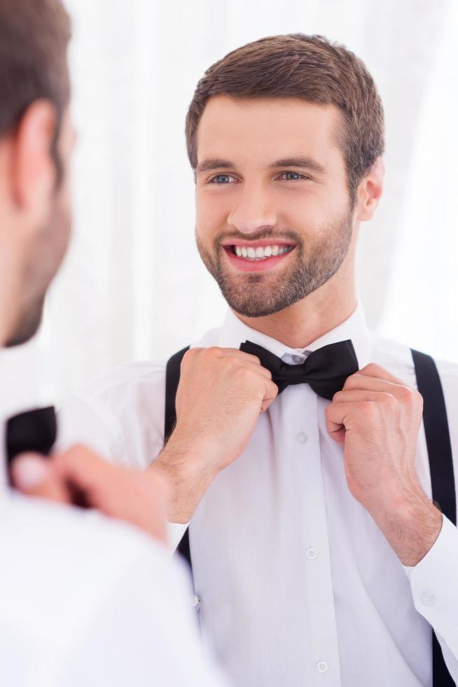 gelukkig bruidegom. detailopname van jong Mens in wit overhemd aanpassen zijn boog stropdas en glimlachen terwijl staand tegen spiegel foto