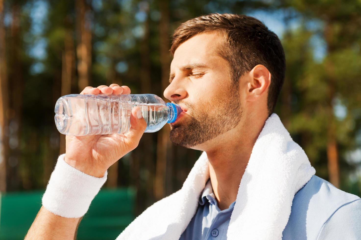 krijgen verfrist na spel. kant visie van dorstig jong Mens in polo overhemd en handdoek Aan schouders drinken water foto