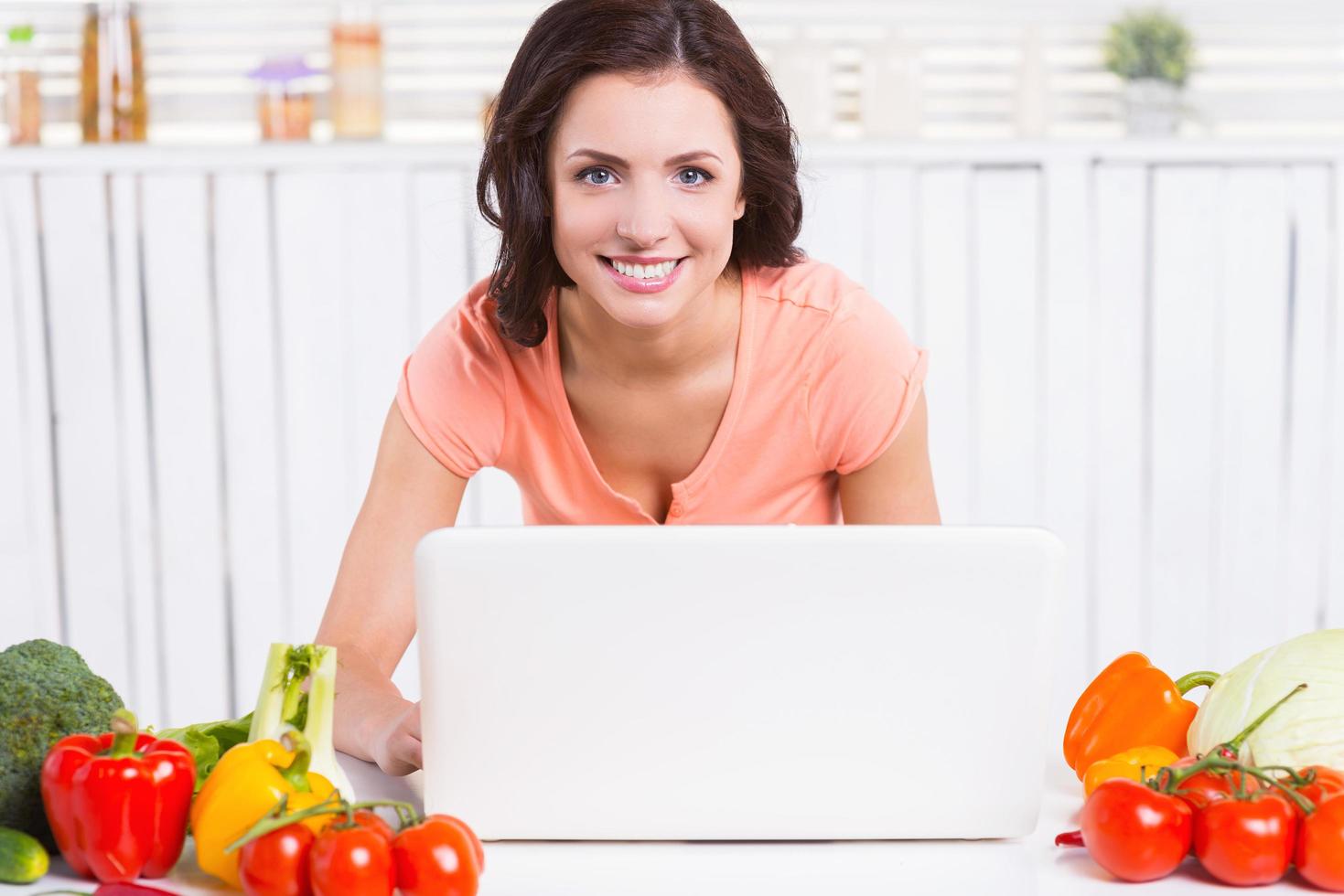 overleg plegen online over Koken. aantrekkelijk jong vrouw gebruik makend van laptop en glimlachen terwijl leunend Bij de keuken tafel met kleurrijk groenten houdende in de buurt haar foto
