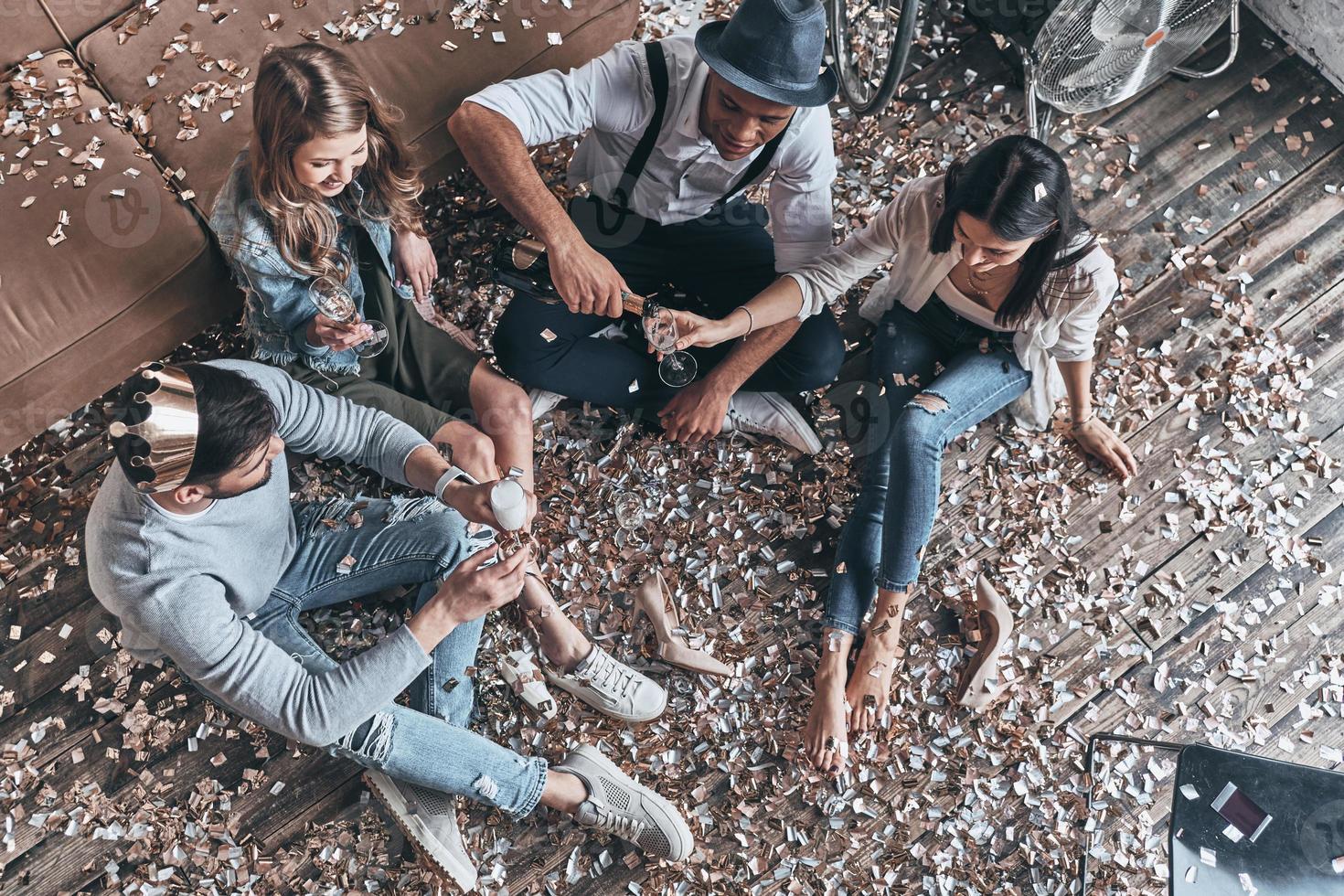 proost naar vriendschap. top visie van modern jong mensen gieten Champagne terwijl zittend Aan de verdieping met confetti in de omgeving van foto