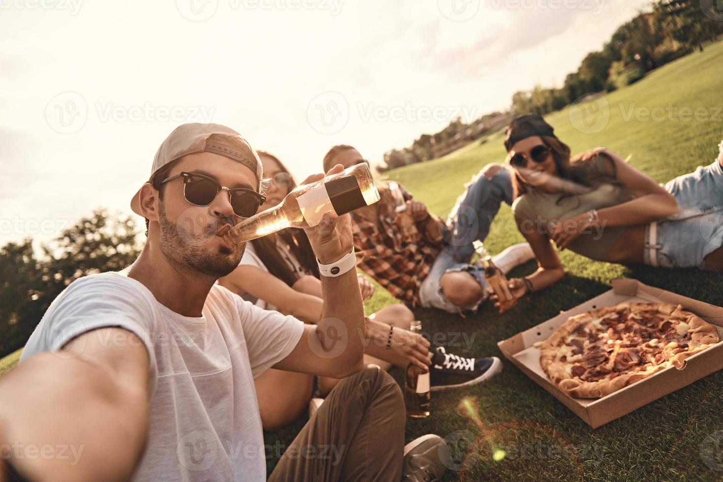 zelf portret van jong mensen in gewoontjes slijtage glimlachen terwijl genieten van pizza en bier buitenshuis foto