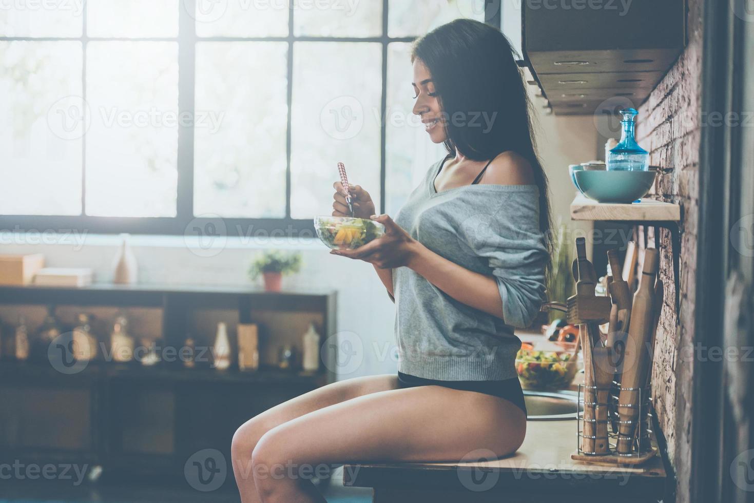 gezond voedsel middelen gezond leven. kant visie van mooi jong gemengd ras vrouw aan het eten salade en glimlachen terwijl zittend Aan de keuken bureau Bij huis foto