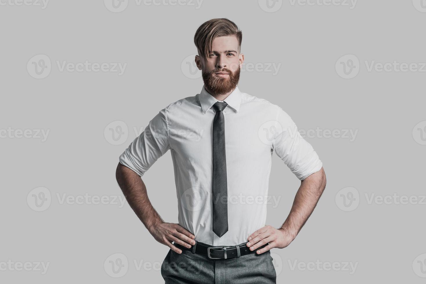 jong en succesvol. knap jong Mens in formele kleding houden handen Aan heup en op zoek Bij camera terwijl staand tegen grijs achtergrond foto