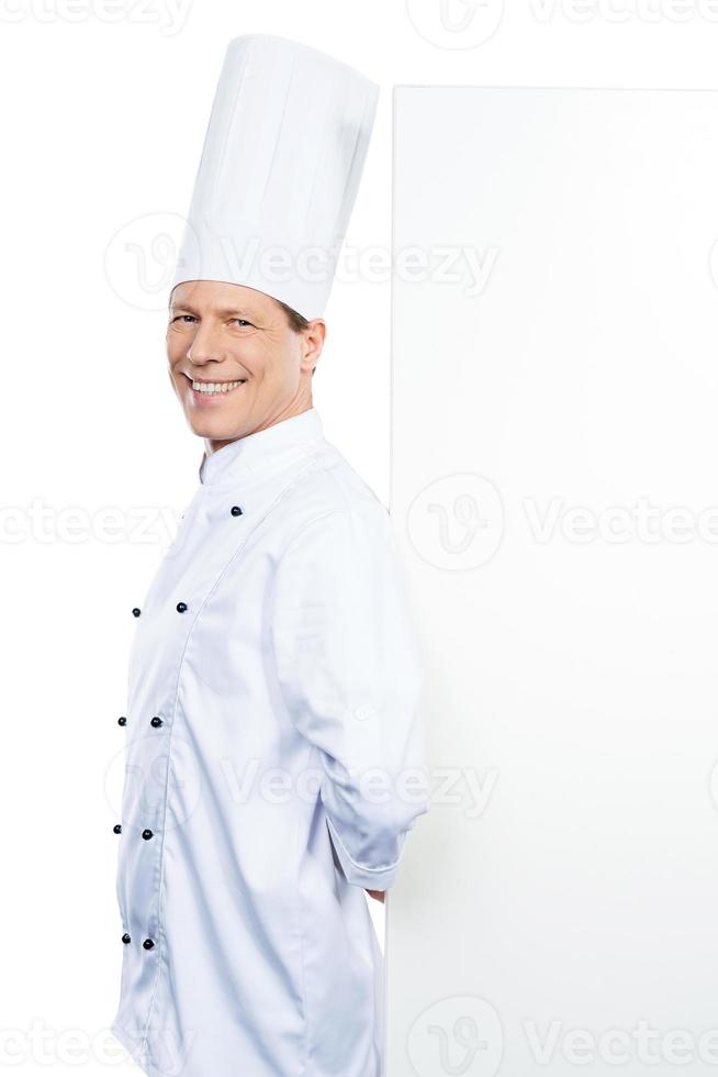 zelfverzekerd en ervaren chef. zelfverzekerd volwassen chef in wit uniform leunend Bij de kopiëren ruimte en glimlachen terwijl staand tegen wit achtergrond foto