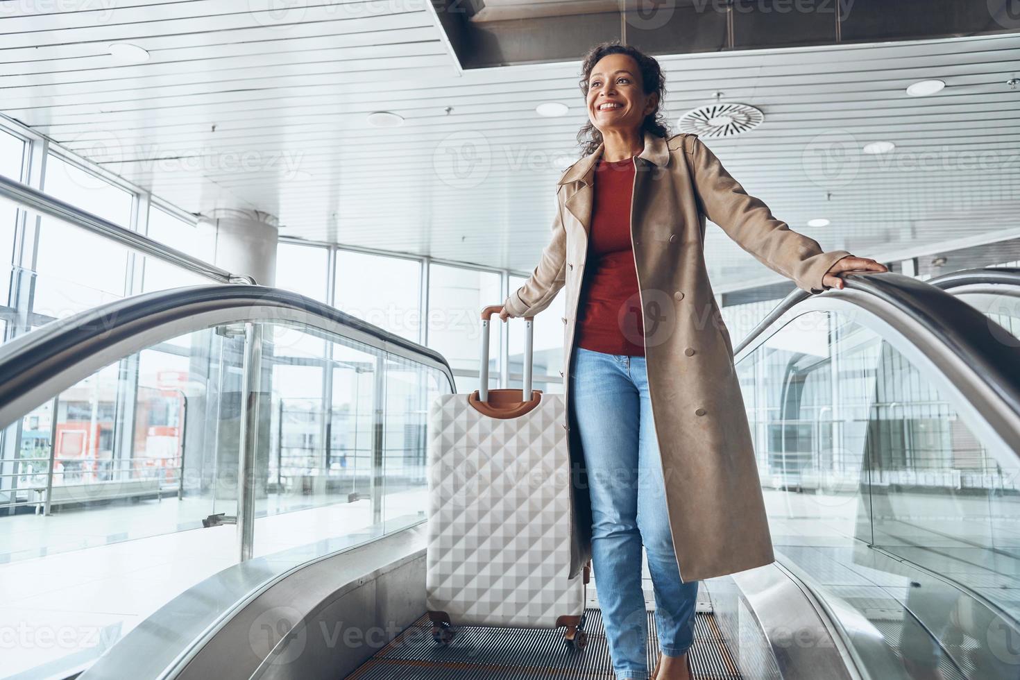 mooi volwassen vrouw glimlachen en op zoek weg terwijl in beweging door roltrap in luchthaven terminal foto