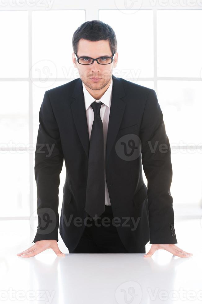 zelfverzekerd zakenman. zelfverzekerd jong Mens in formele kleding op zoek Bij camera terwijl leunend Bij de tafel foto