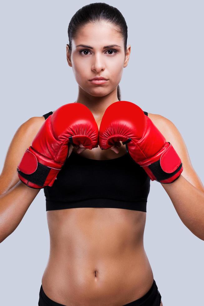 energie binnen haar. aantrekkelijk jong sportief vrouw in boksen handschoenen op zoek kunst camera terwijl staand tegen grijs achtergrond foto