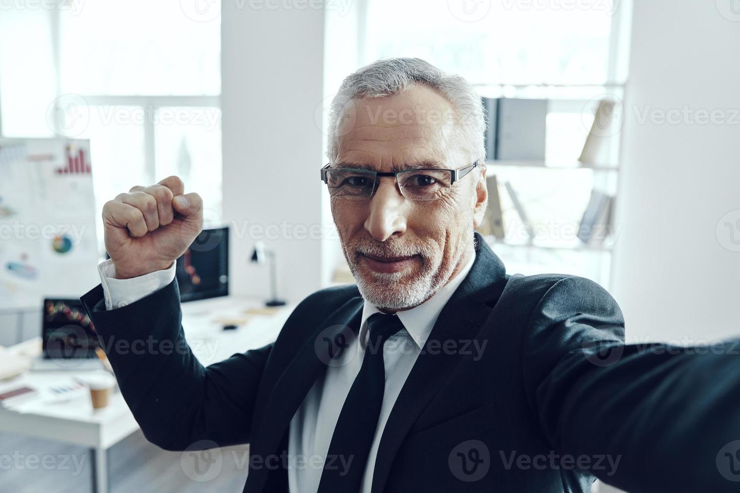 zelf portret van senior Mens in elegant bedrijf pak op zoek Bij camera en glimlachen terwijl staand in kantoor foto