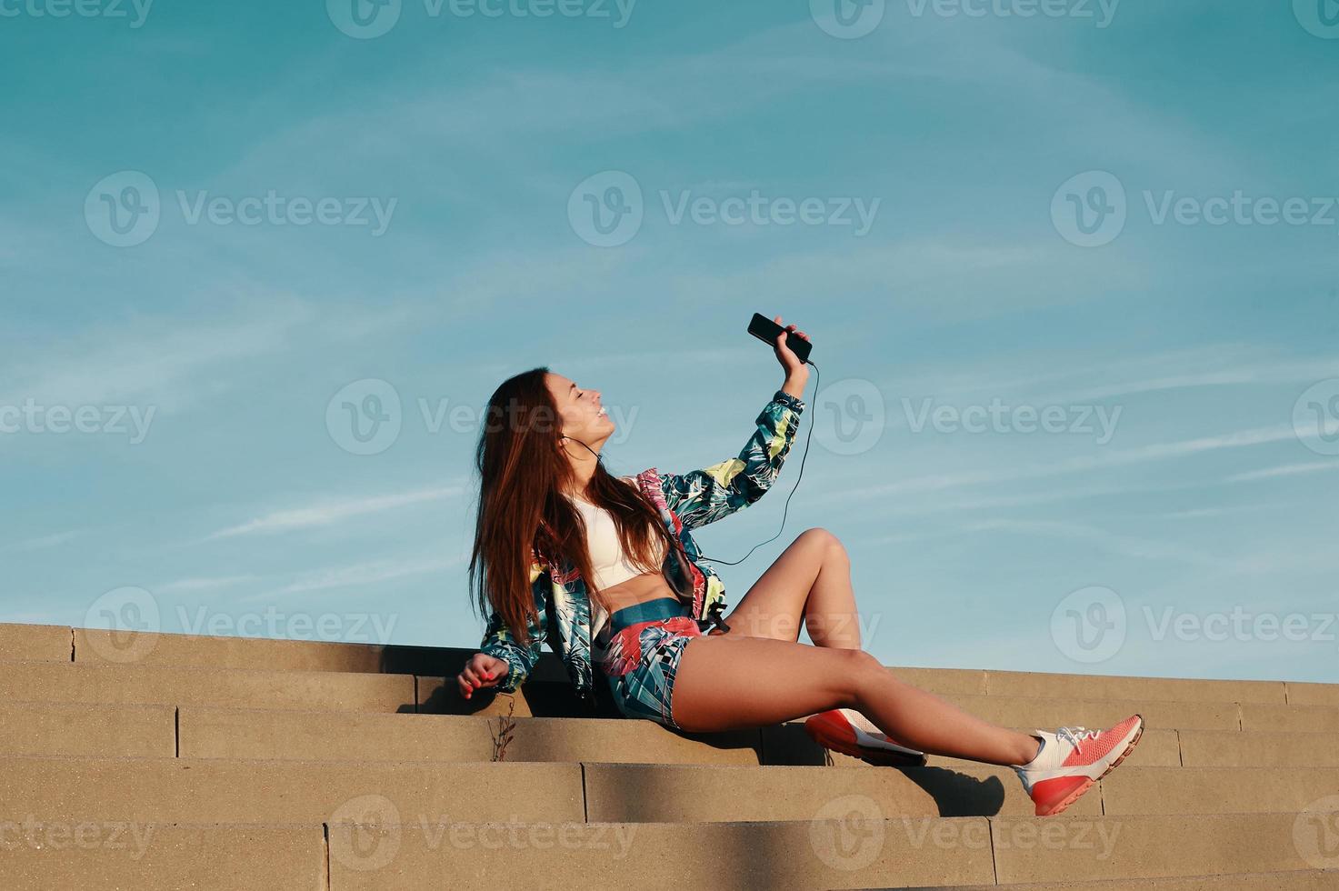 luisteren naar favoriete muziek. aantrekkelijk jong vrouw in sport- kleding glimlachen terwijl zittend Aan de stappen buitenshuis foto