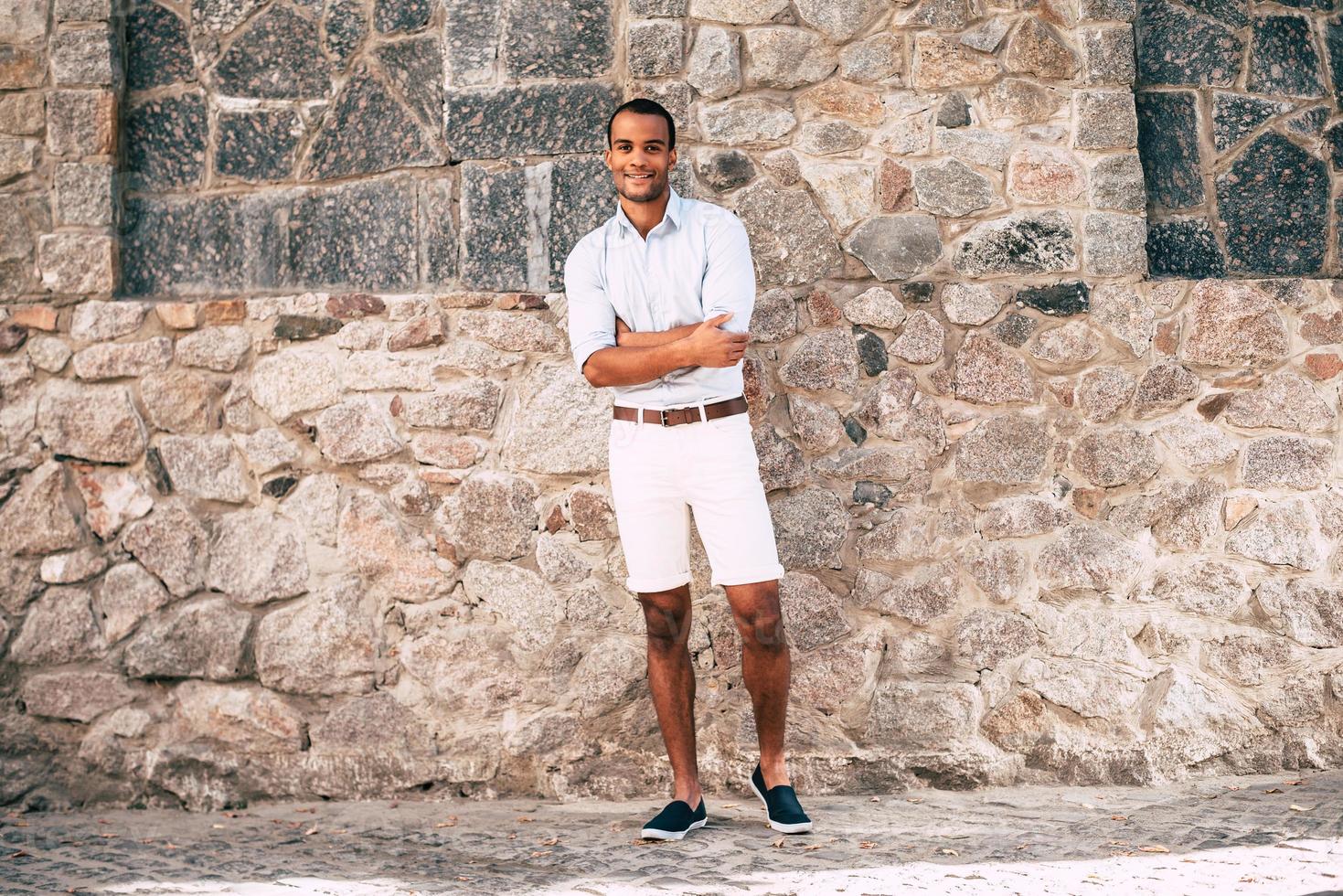 zelfverzekerd en knap. vol lengte van vrolijk jong Afrikaanse Mens in slim gewoontjes kleren houden armen gekruiste en op zoek Bij camera terwijl staand tegen de stoned muur buitenshuis foto