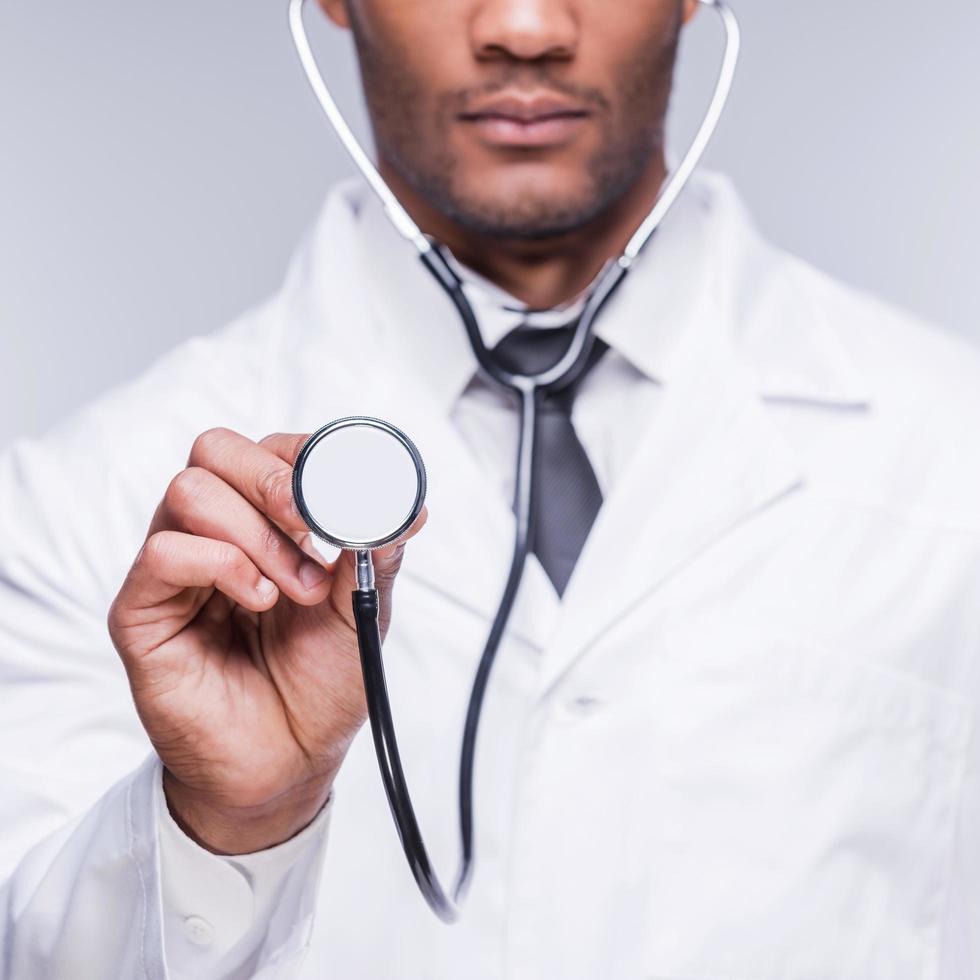 medisch examen. bijgesneden beeld van zelfverzekerd Afrikaanse dokter uitrekken uit zijn stethoscoop terwijl staand tegen grijs achtergrond foto