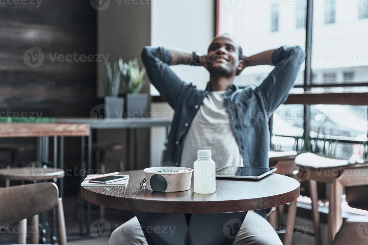 gevoel ontspannen. vrolijk jong Afrikaanse Mens houden handen achter hoofd terwijl zittend in de cafetaria foto