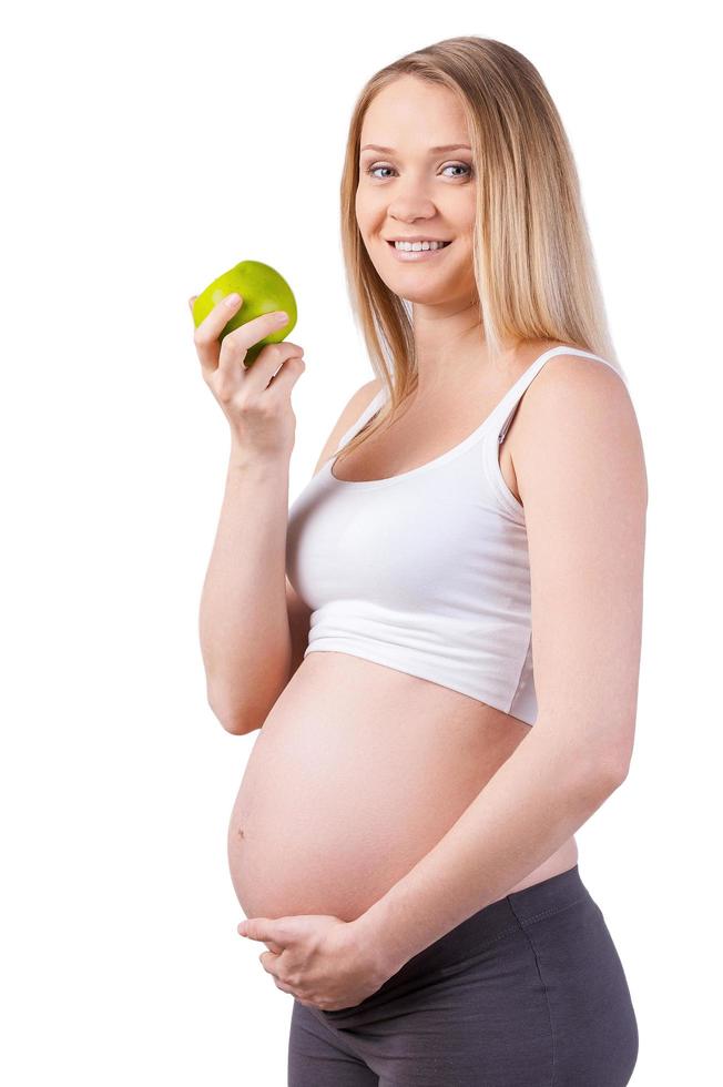 aan het eten enkel en alleen gezond voedsel. vrolijk zwanger vrouw Holding groen appel en op zoek Bij camera terwijl staand geïsoleerd Aan wit foto