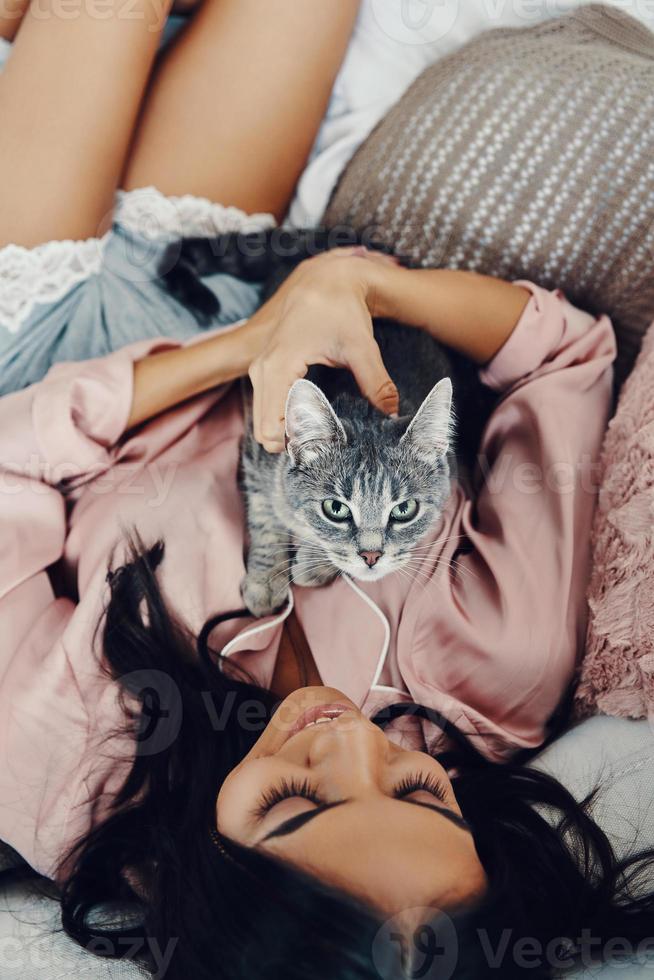 top visie van mooi jong vrouw in pyjama glimlachen en omarmen huiselijk kat terwijl resting in bed Bij huis foto