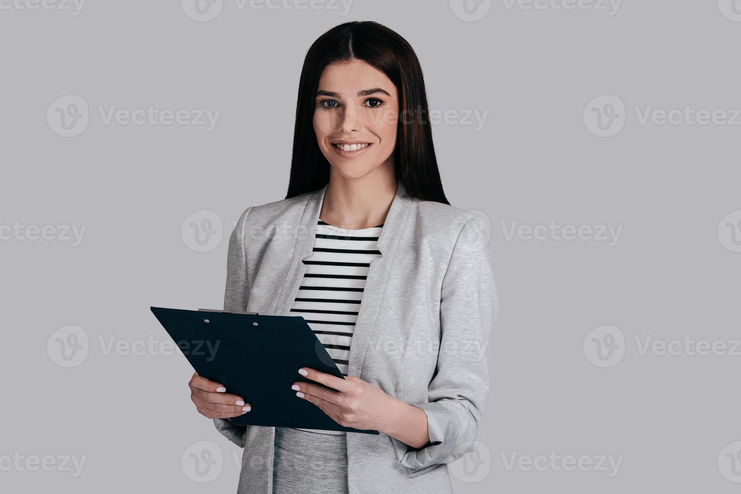 altijd klaar naar werk. mooi jong vrouw in slim gewoontjes slijtage glimlachen en op zoek Bij camera terwijl staand tegen grijs achtergrond foto