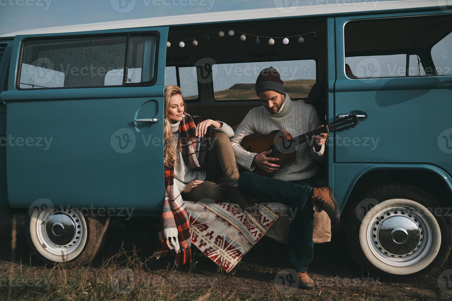 spelen haar favoriete liedje. knap jong Mens spelen gitaar voor zijn mooi vriendin terwijl zittend in blauw retro stijl mini busje foto