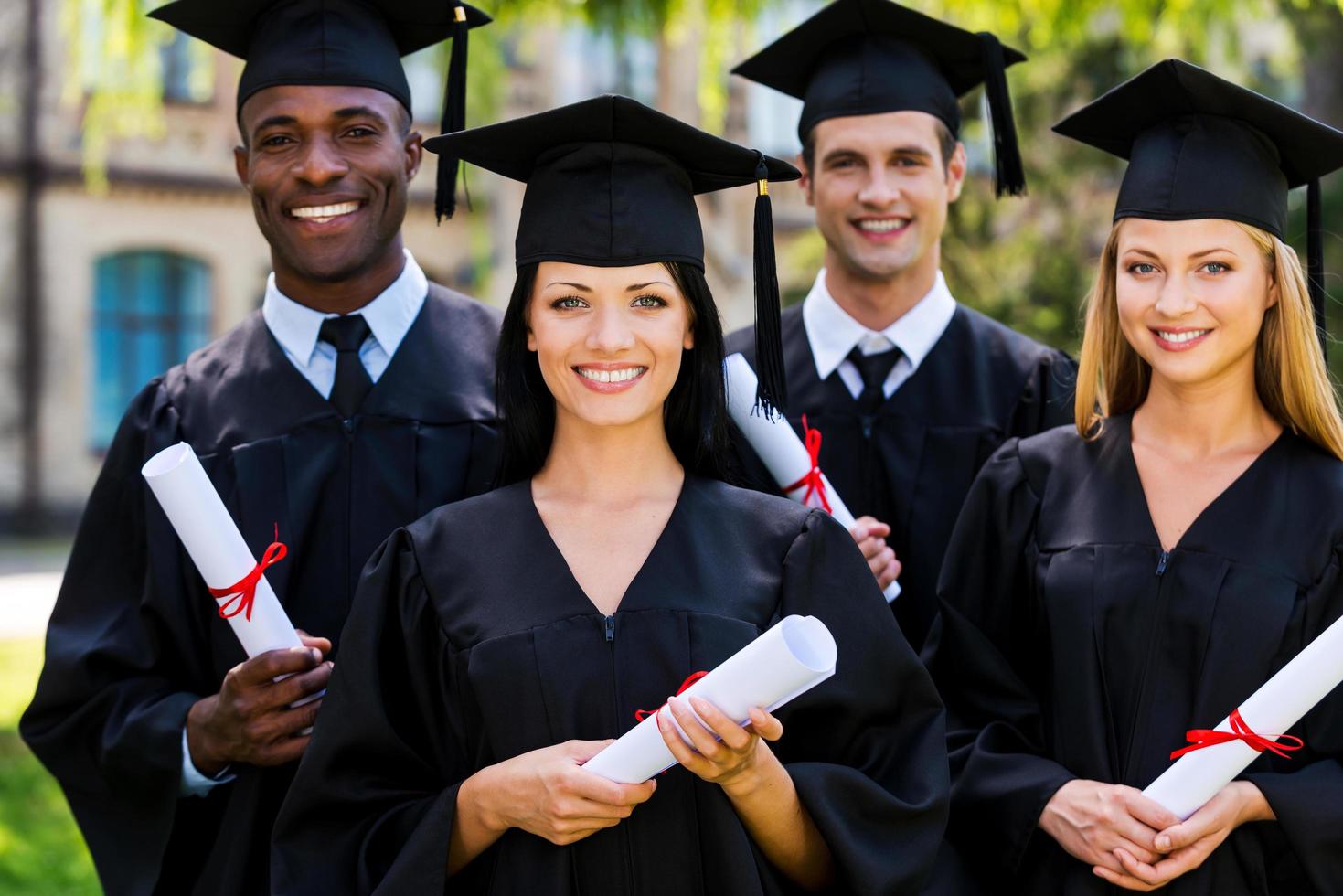 gevoel zelfverzekerd in hun toekomst. vier college afgestudeerden in diploma uitreiking jurken staand dichtbij naar elk andere en glimlachen foto