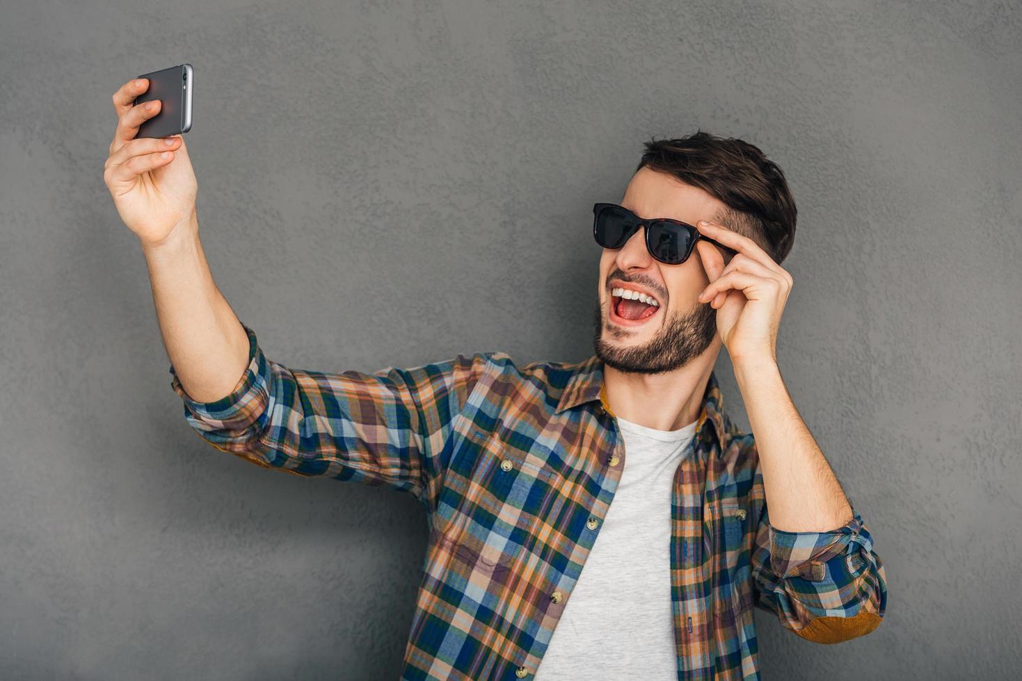 tijd voor selfie. vrolijk jong Mens in zonnebril maken selfie terwijl staand tegen grijs achtergrond foto