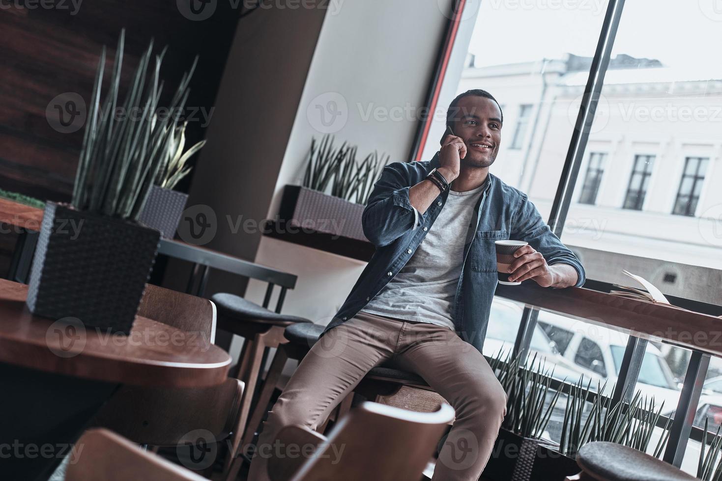 planning weekend met zijn vriendin. knap jong Mens pratend Aan zijn slim telefoon met glimlach terwijl zittend in de cafetaria foto