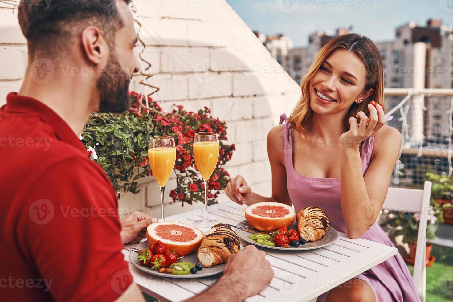 romantisch jong paar in gewoontjes kleding hebben ontbijt en glimlachen terwijl zittend Aan de op het dak patio buitenshuis foto