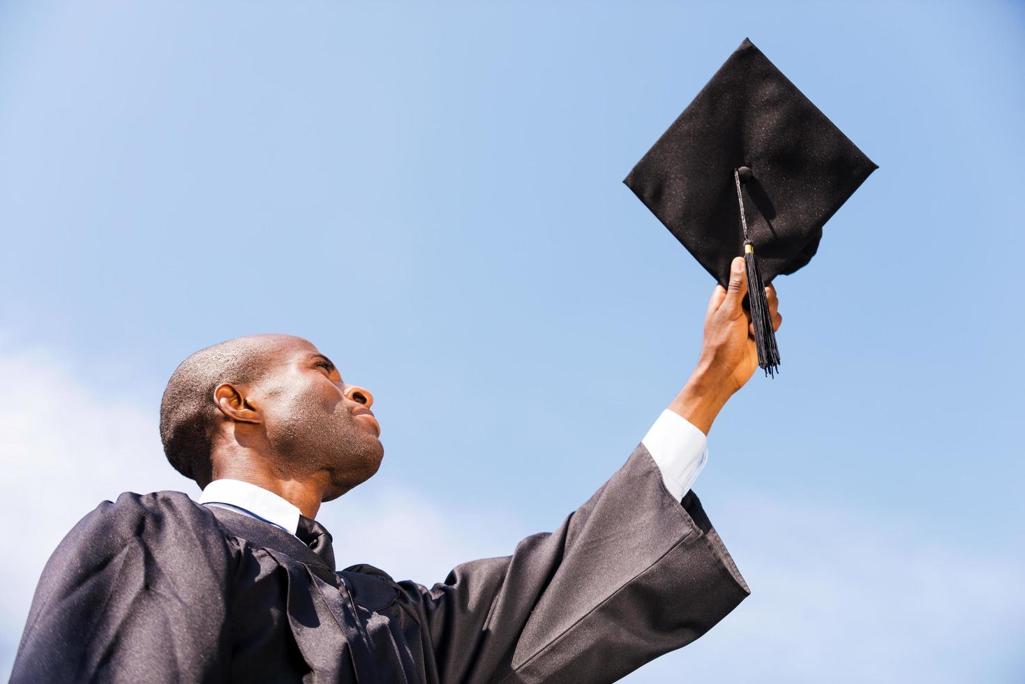 gelukkig afstuderen. laag hoek visie van gelukkig jong Afrikaanse Mens in diploma uitreiking japon Holding zijn Mortier bord tegen blauw lucht foto