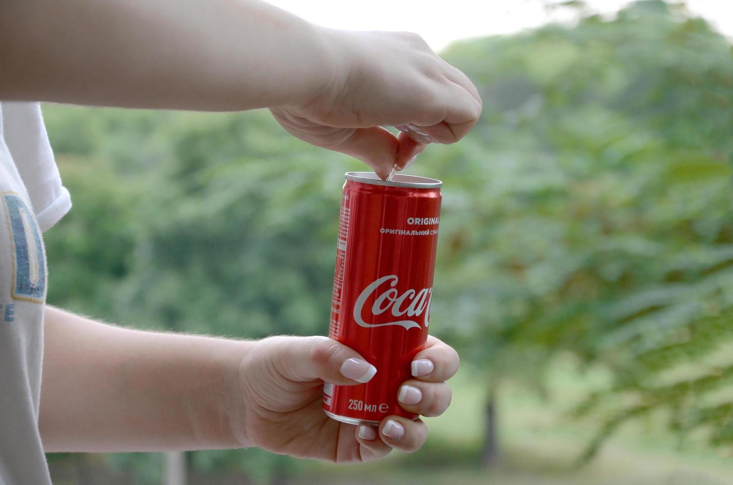 Charkov. Oekraïne - mei 2, 2019 Kaukasisch vrouw houdt rood Coca Cola blik kan met groen tuin achtergrond foto