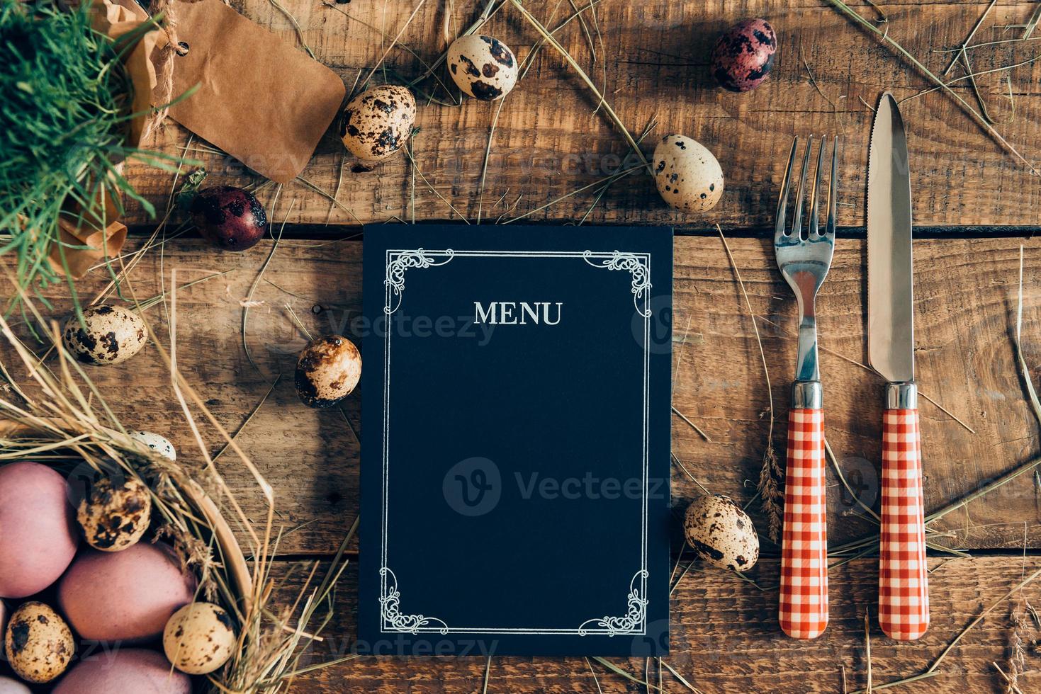 Pasen avondeten menu. top visie van Pasen eieren en menu bord met vork en mes aan het liegen Aan houten rustiek tafel met hooi foto