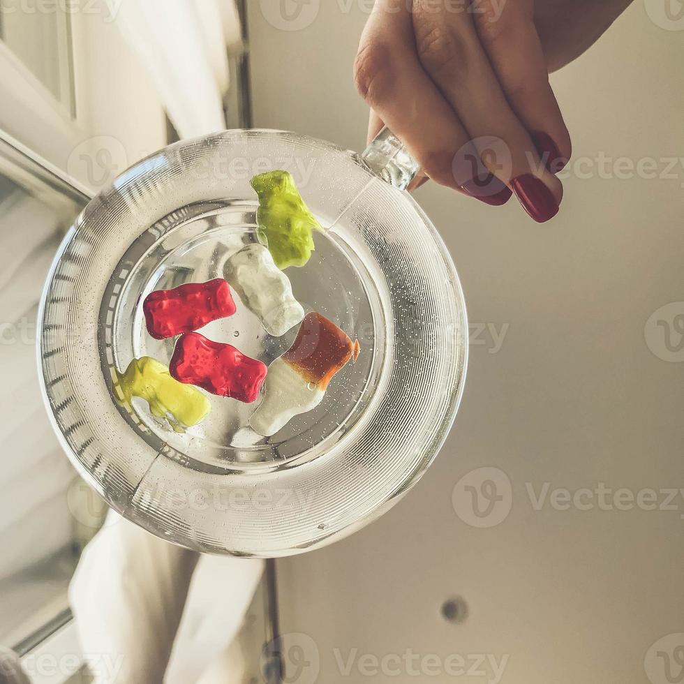 transparant mok, ronde en groot, gevulde met water. Bij de bodem van de mok zijn kleverig bears en flessen van limonade. meisje met helder rood manicure houdt een mok in haar handen. Aan wit matte achtergrond foto