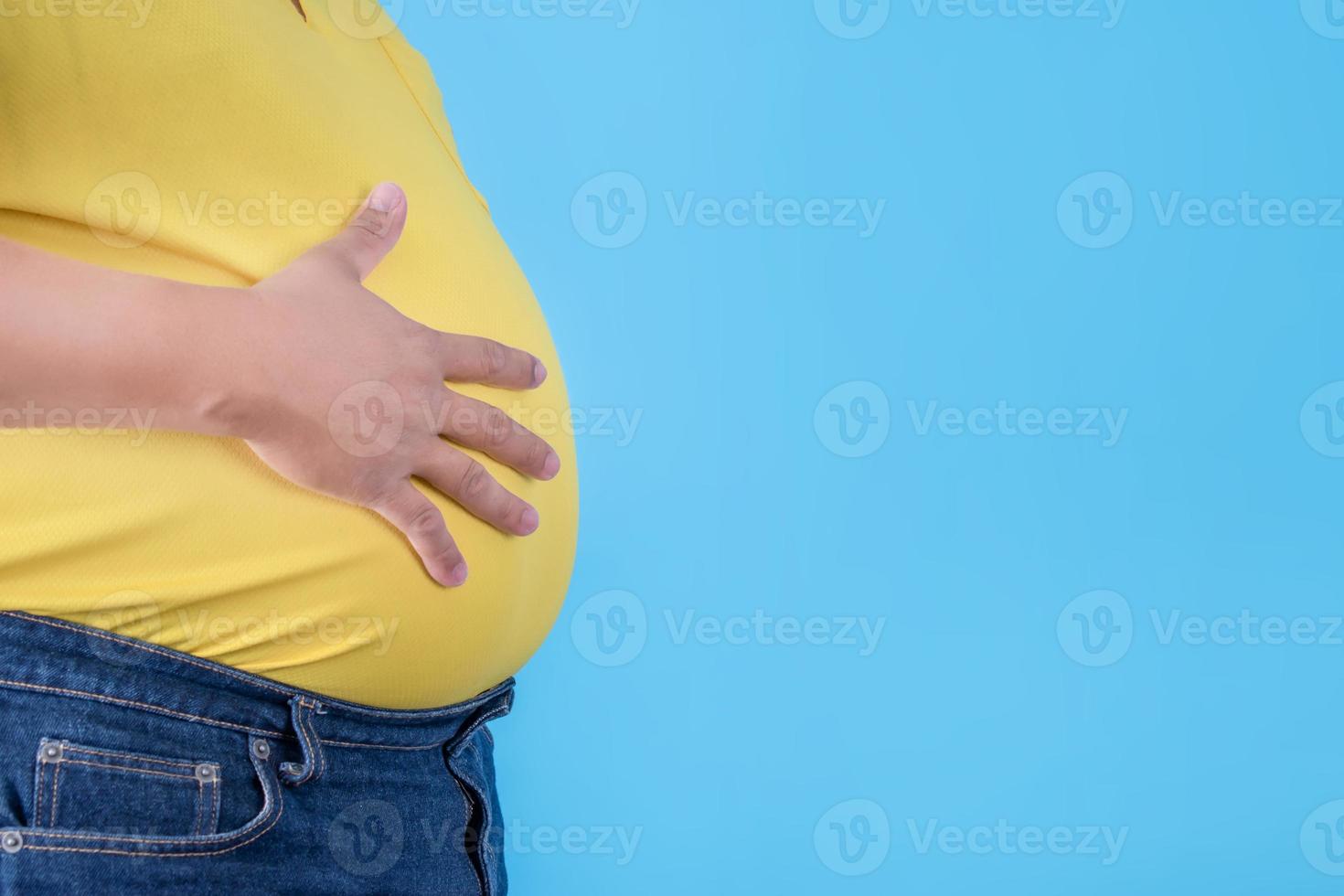 de maag van een te zwaar Mens is vervelend een geel overhemd en jeans Aan een blauw achtergrond. foto