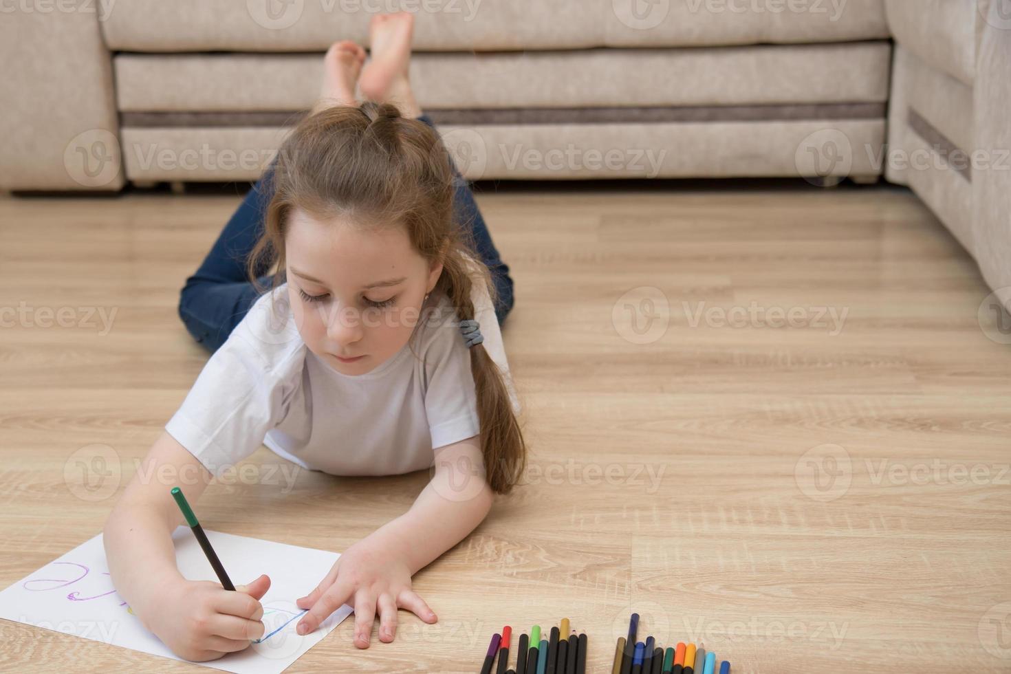 schattig weinig meisje in jeans leugens Aan de verdieping Bij huis en trekt met gekleurde potloden, de concept van de ontwikkeling van een kleuter, hobby's foto