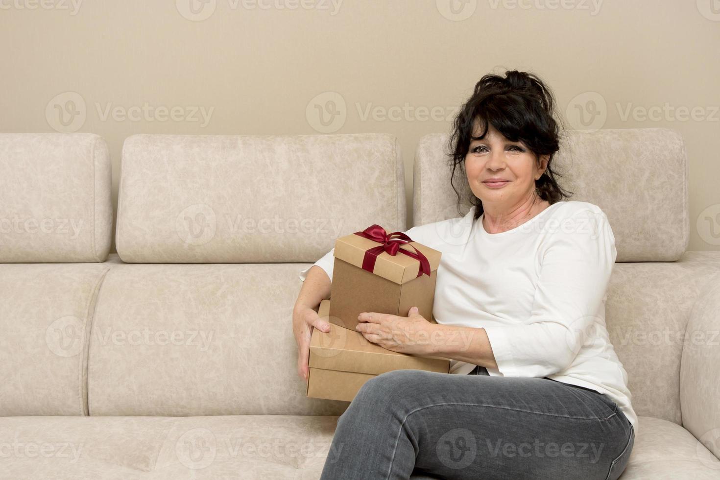 mooi oud vrouw zit Aan de sofa met cadeaus in haar handen en glimlacht foto