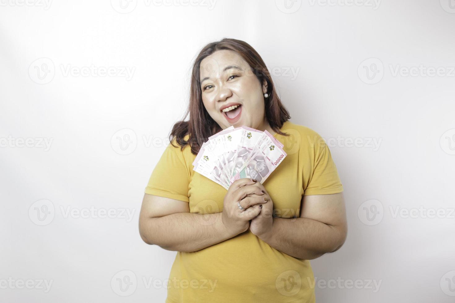 een gelukkig jong groot grootte vrouw is vervelend geel t-shirt en Holding contant geld geld in Indonesisch roepia geïsoleerd door wit achtergrond foto