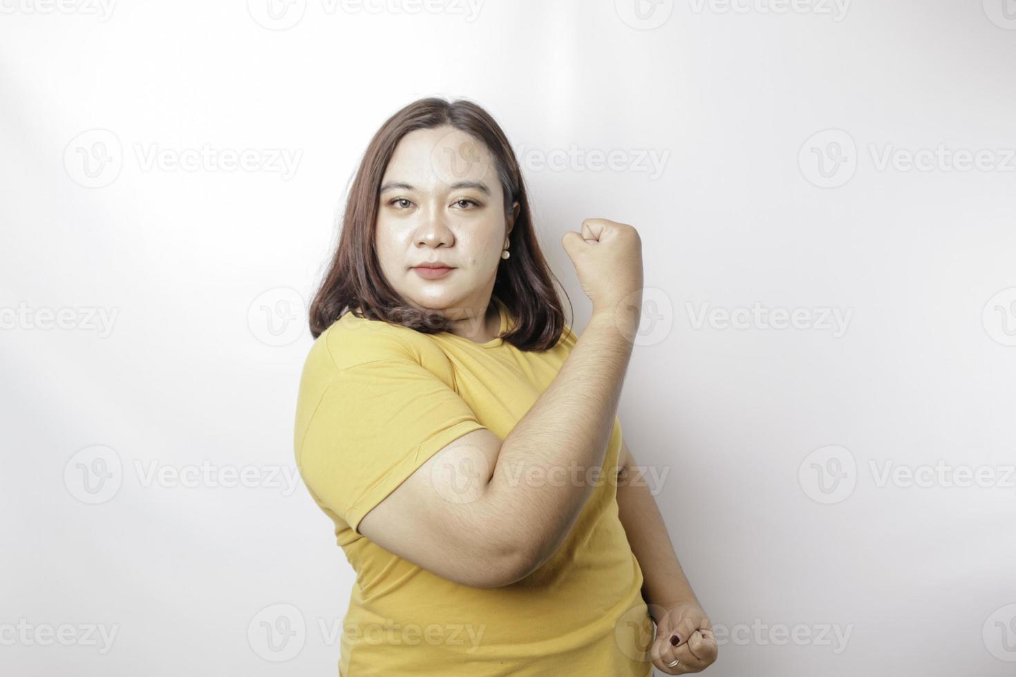 opgewonden Aziatisch groot formaat vrouw vervelend een geel t-shirt tonen sterk gebaar door hijs- haar armen en spieren glimlachen trots foto