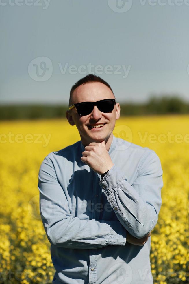 een jong Mens in zonnebril is wandelen en hebben pret door een zonnig geel koolzaad veld, de concept van reizen en vrijheid. zomer vakantie achtergrond foto