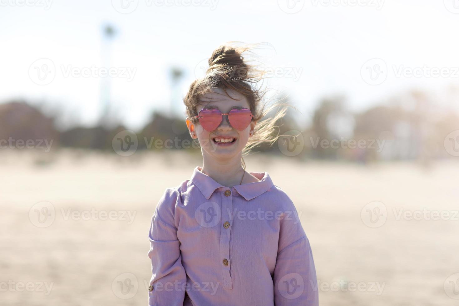 glimlachen gelukkig weinig meisje in zonnebril in het formulier hart is hebben pret Aan strand. zomer vakantie vakantie. kind is op zoek Aan camera Aan palm bomen achtergrond foto