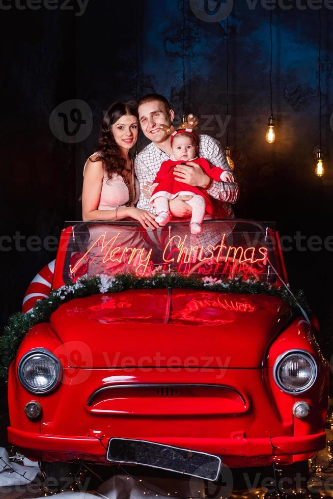 mama, vader en baby meisje hebben pret in de Kerstmis retro rood auto. ouders met een weinig dochter in een Kerstmis foto sessie. studio het schieten voordat de vakantie.